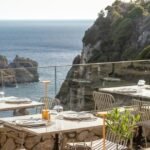 Seaside view of Paleokastritsa bay in Corfu with tavernas overlooking turquoise water and boats in the distance.