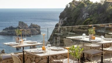Seaside view of Paleokastritsa bay in Corfu with tavernas overlooking turquoise water and boats in the distance.